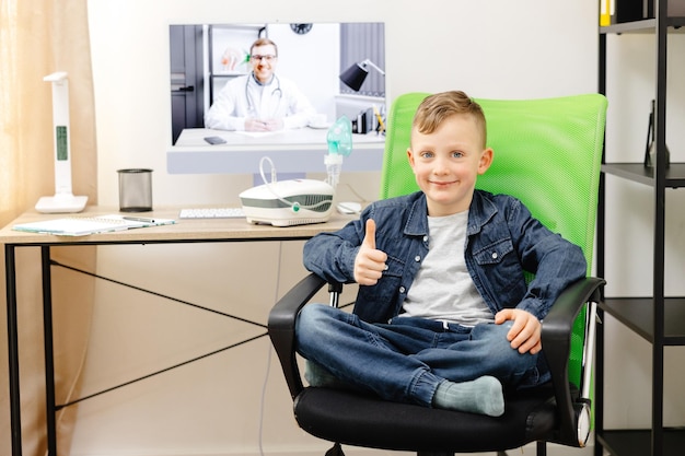 Foto un lindo niño sonriente está sentado en una silla en casa y en el fondo hay una mesa con una computadora en la que un médico está realizando una consulta en línea