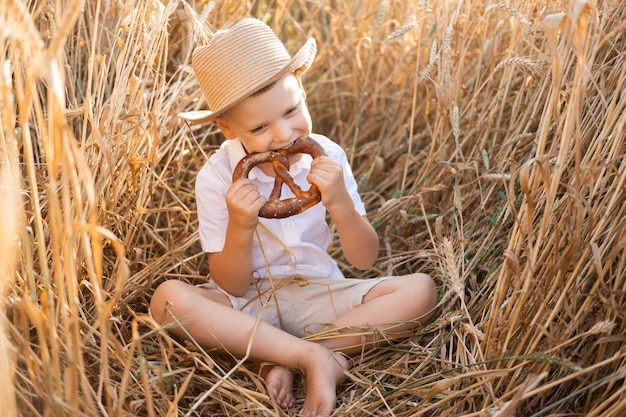 lindo niño sonriente con sombrero de paja comiendo brezel en el campo de trigo al atardecer de verano