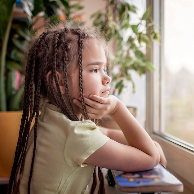 Lindo niño triste sentado en el alféizar de la ventana y mirando a la calle