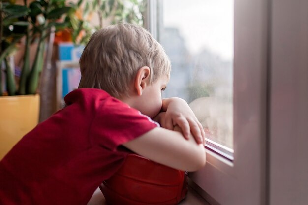 Lindo niño triste sentado en el alféizar de la ventana y mirando a la calle