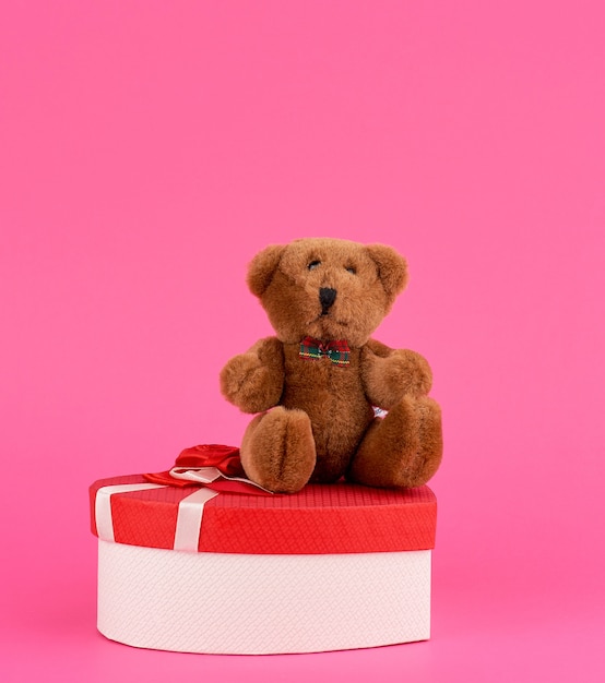 Lindo oso de peluche marrón y caja de regalo roja sobre un fondo rosa