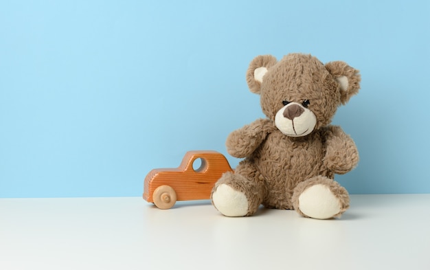 Lindo oso de peluche marrón se sienta en una mesa blanca y un coche de juguete para niños de madera, fondo azul.