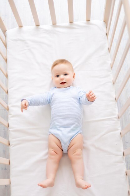 Un lindo y saludable bebé está acostado boca arriba en una cuna sobre ropa de cama blanca en casa con un traje azul. El niño feliz mira a la cámara y sonríe.