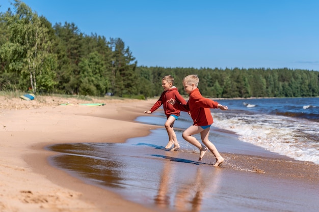 Lindos muchachos caucásicos vistiendo sudaderas con capucha rojas y calzoncillos azules corriendo de las olas en el lago Ladoga