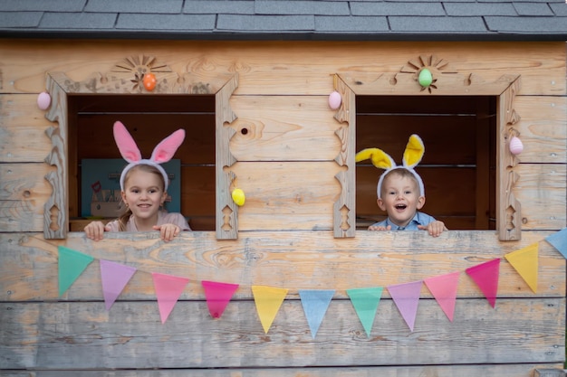 Lindos niños alegres con orejas de conejo en la casa de juegos celebran la pascua hermano y hermana mirando por la ventana