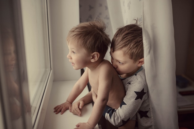 Lindos niños caucásicos mirando por la ventana un niño besando el hombro de su hermano pequeño