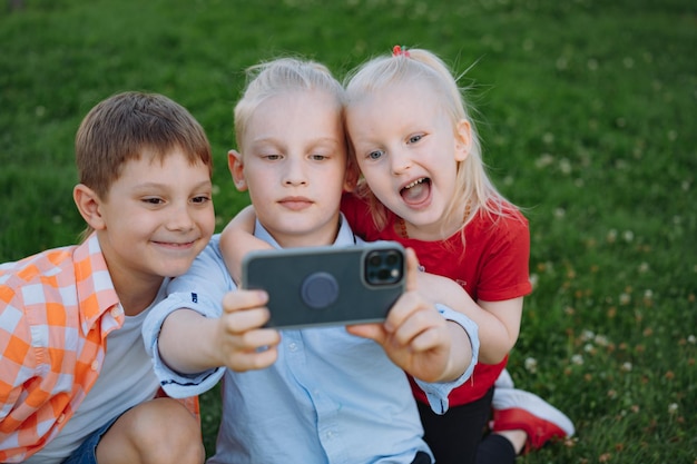 lindos niños divertidos disparando selfie con teléfono inteligente. Niños en el parque sentados en el césped divirtiéndose juntos. Imagen con enfoque selectivo