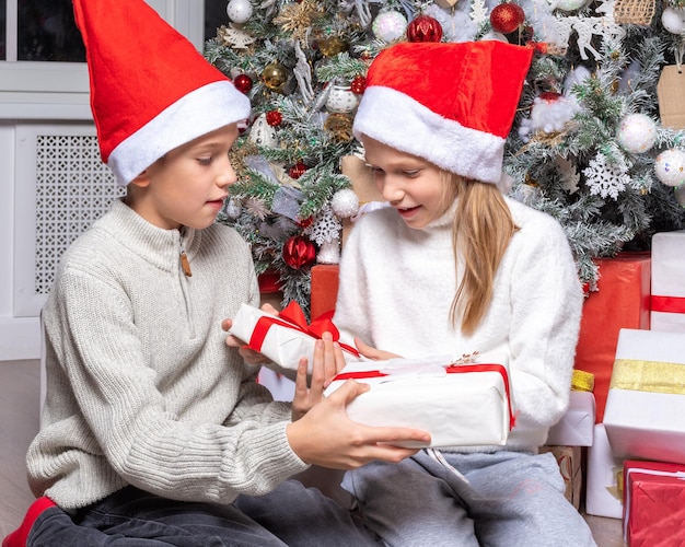 Lindos niños felices emocionados niño y niña con sombreros de santa intercambian regalos sorpresa junto al árbol de navidad en casa