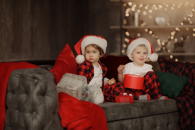 Lindos niños con gorro de Papá Noel sentado en una gran silla acogedora en la habitación con caja de regalos de Navidad