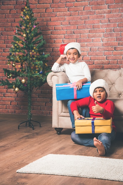 Lindos niños indios asiáticos celebrando la Navidad en casa con gorro de Papá Noel, regalos y árbol de Navidad