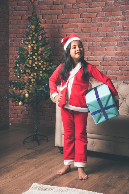 Lindos niños indios asiáticos celebrando la Navidad en casa con gorro de Papá Noel, regalos y árbol de Navidad