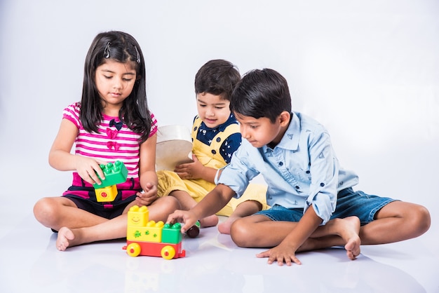 Lindos niños indios o asiáticos jugando con juguetes o bloques y divirtiéndose mientras está sentado en la mesa o aislado sobre fondo blanco.