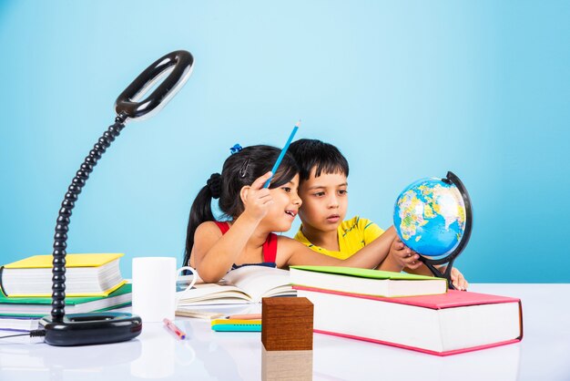 Lindos niños indios o asiáticos que estudian en la mesa de estudio con pila de libros, globo educativo, aislado sobre color azul claro