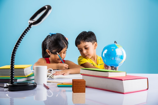 Lindos niños indios o asiáticos que estudian en la mesa de estudio con pila de libros, globo educativo, aislado sobre color azul claro