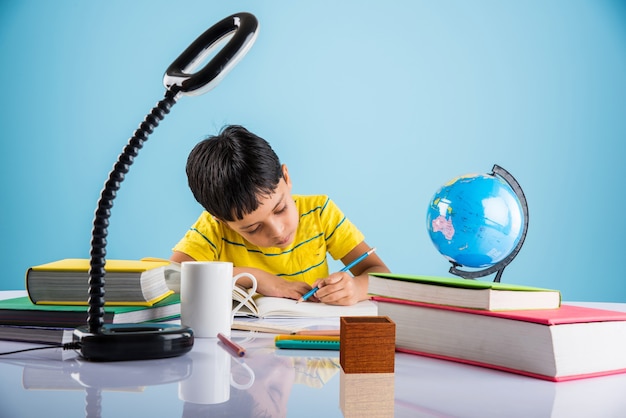 Lindos niños indios o asiáticos que estudian en la mesa de estudio con pila de libros, globo educativo, aislado sobre color azul claro