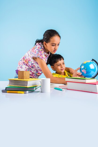 Lindos niños indios o asiáticos que estudian en la mesa de estudio con pila de libros, globo educativo, aislado sobre color azul claro