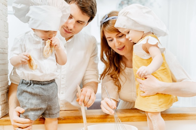 Lindos padres positivos, mamá y papá, enseñan a sus adorables gemelos y una niña con trajes de cocinera cómo amasar la masa. El concepto de clases conjuntas para niños y padres.