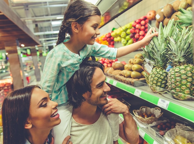 Foto lindos pais jovens e sua filha bonitinha estão conversando e sorrindo enquanto escolhem comida no supermercado a menina está sentada nos ombros do pai