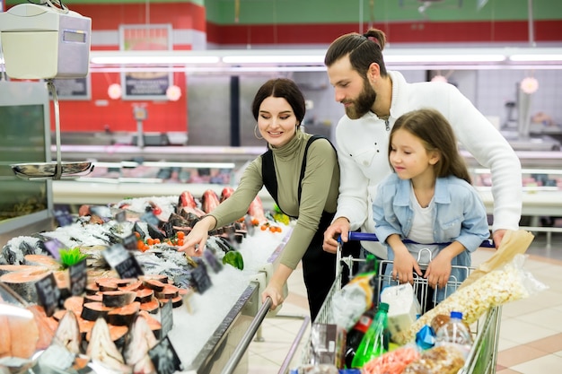 Lindos pais jovens e sua filhinha estão sorrindo enquanto escolhem peixes no supermercado
