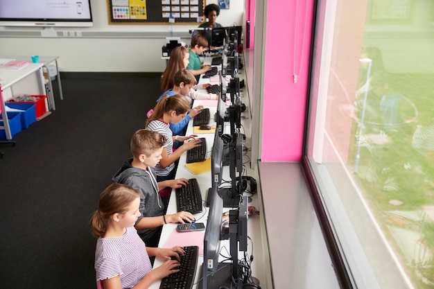 Línea de estudiantes de secundaria trabajando en pantallas en clase de computación con profesora en segundo plano tomada desde un ángulo alto