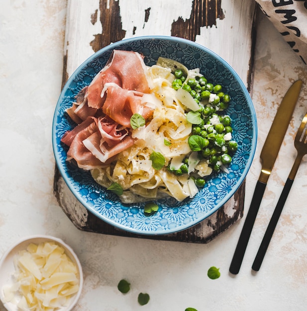 Linguine pasta con salsa blanca, guisantes verdes, parmesano, jamón y menta fresca en un plato azul sobre tablero rústico.