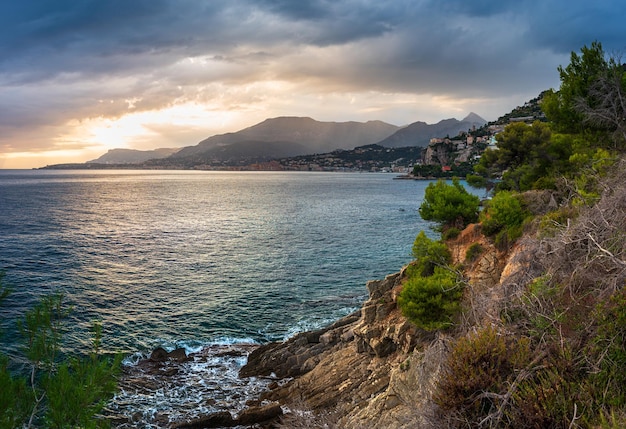 Linha da costa rochosa e baía de água no inverno, riviera dei fiori, liguria, itália. cidade de menton em segundo plano. céu dramático ao pôr do sol.