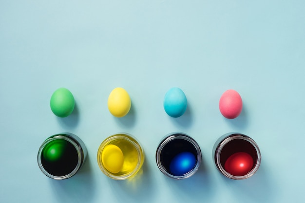 Foto linha de ovos de páscoa coloridos e potes cheios de tintura de ovo em fundo azul pastel. postura plana.