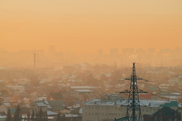 Linhas de energia na cidade ao amanhecer. silhuetas de edifícios urbanos entre poluição atmosférica no nascer do sol. cabos de alta tensão no céu amarelo laranja quente. indústria de energia ao pôr do sol. fonte de alimentação da cidade. meio urbano de névoa.