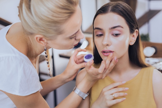 Lippen des Kunden. Professionelle geschickte blonde Maskenbildnerin, die ihre Arbeit genießt, während Lippen des Klienten färbt