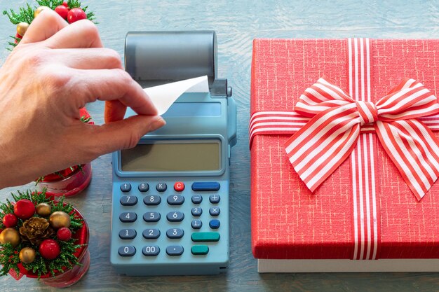 Liquidação de Natal. Mão segurando um cheque em branco em uma caixa registradora perto de uma caixa de presente vermelha e árvores de Natal