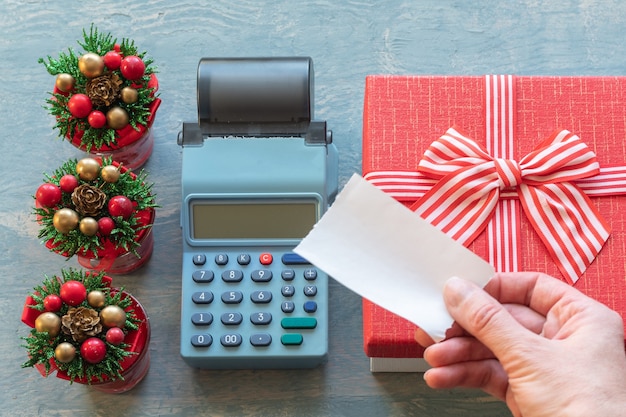 Liquidação de Natal. Uma mão segurando um cheque em branco acima da caixa registradora perto de uma caixa de presente vermelha com uma fita e pequenas árvores de Natal com cones. Compras online.