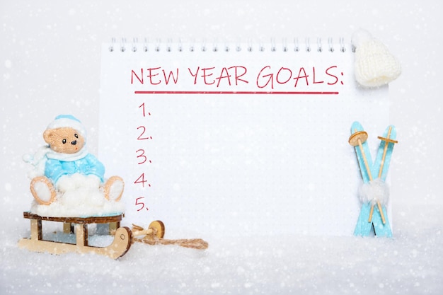 Lista de metas para o feriado de Ano Novo. Urso de pelúcia com roupas azuis sentado em um trenó, esquis de madeira azuis, um chapéu branco e um caderno em branco com as palavras OBJETIVOS DE ANO NOVO na neve branca