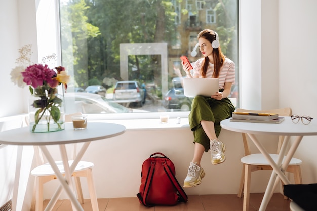 Lista de reprodução favorita. Mulher séria segurando laptop sobre os joelhos enquanto está sentada no parapeito da janela