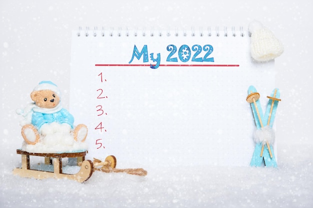 Lista de metas de año nuevo. Oso de peluche con ropa azul sentado en un trineo, esquís de madera azul, un sombrero blanco y un cuaderno con la inscripción MY 2022 sobre un fondo de nieve blanca. Concepto de deporte de invierno.