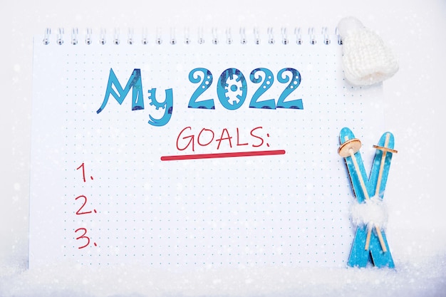 Liste der Ziele für die Neujahrsfeiertage. Hellblaue Holzskier und ein Notizbuch mit der Aufschrift MY 2022 GOALS auf weißem Schnee. Wintersportkonzept. Sportziele für das Neujahrskonzept