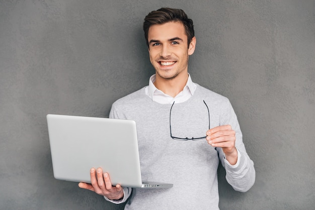 Foto listo para ayudarte. hombre joven confidente con sonrisa y sosteniendo la computadora portátil mientras está de pie