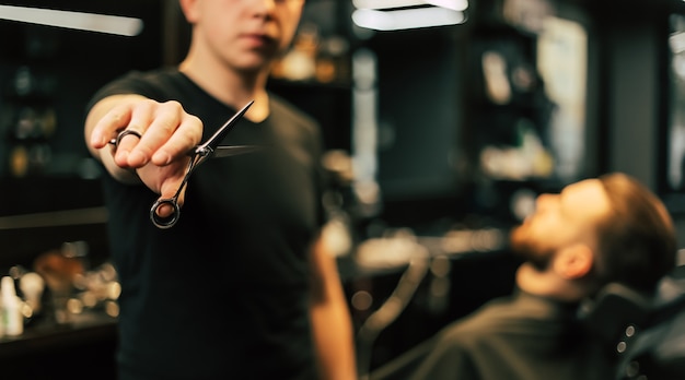 Listo para darle estilo. Foto de primer plano de un joven peluquero con una camiseta negra que sostiene unas tijeras en su mano derecha estirada hacia la cámara.