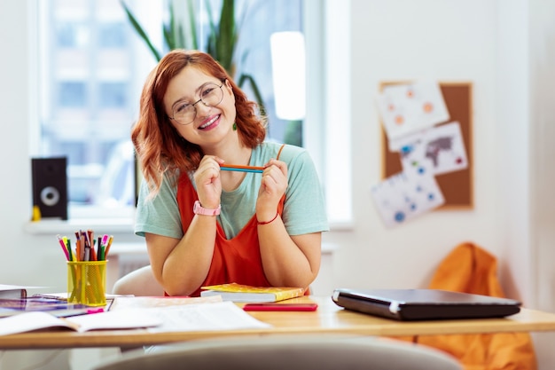 Listo para estudiar. Bonita mujer alegre sonriendo mientras está sentado en el escritorio