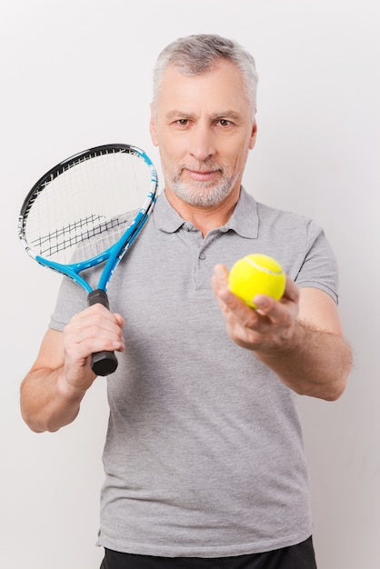 ¿Listo para jugar? Hombre mayor de pelo gris confiado que sostiene la raqueta de tenis y que le da una pelota de tenis mientras está de pie contra el fondo blanco