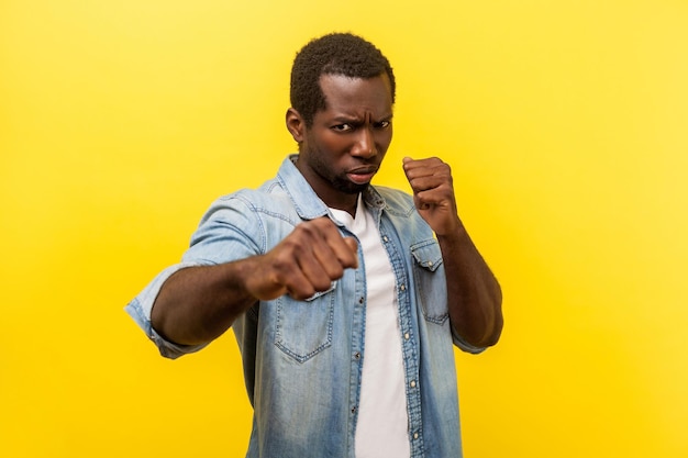 Listo para la pelea. Retrato de hombre agresivo en camisa de denim con mangas enrolladas de pie en postura defensiva con puños apretados, amenazando con atacar. tiro de estudio interior aislado en amarillo