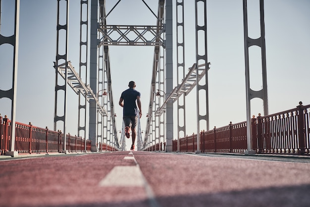 Listo para romper su récord. Vista trasera de la longitud completa del joven africano en ropa deportiva haciendo ejercicio mientras trota en el puente al aire libre