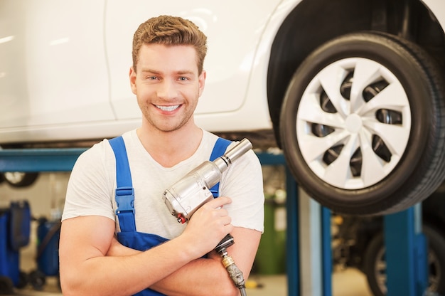 Listo para trabajar. Hombre joven confidente que sostiene una llave neumática y sonriendo mientras está de pie en el taller con el coche en el fondo