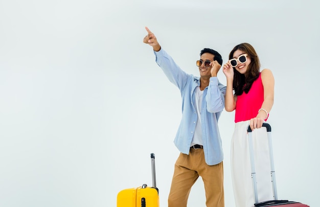 Listo para viajar felices vacaciones Vacaciones de verano Pareja asiática en gafas de sol con señalar y mirar hacia adelante con emocionante joven y hombre con maleta en viaje aislado sobre fondo blanco.