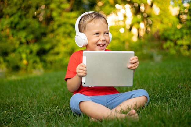 Little Baby Boy en auriculares inalámbricos sosteniendo una tableta al aire libre en un parque de verano sonriendo