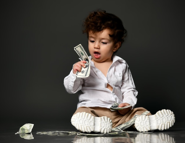 Little Baby-Chef mit Geld Kind-Modell-Kleinkind tragen Freizeitkleidung Schuhe halten Papier-Dollar-Banknote Isolierte Porträt isoliert Schuss auf grauem Studio-Hintergrund Kinder und Finanzen