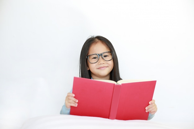 Livro de capa dura asiático pequeno feliz bonito da leitura da menina da criança que encontra-se na cama contra o fundo branco.