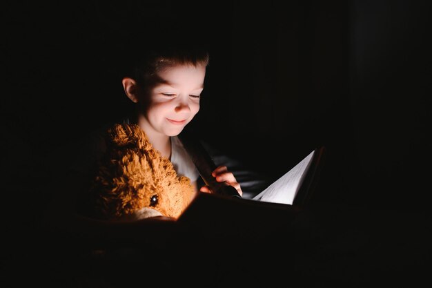 Livro de leitura infantil na cama. As crianças lêem à noite. Menino com livros de conto de fadas no quarto. Educação para crianças pequenas. História de ninar à noite. Garoto bonito sob o cobertor no quarto escuro com lâmpada.