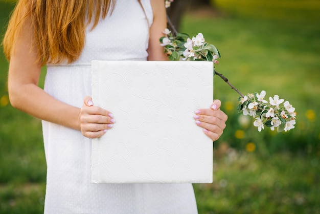 Livro fotográfico em capa de couro branco nas mãos de uma mulher na primavera perto de um galho florido de uma macieira