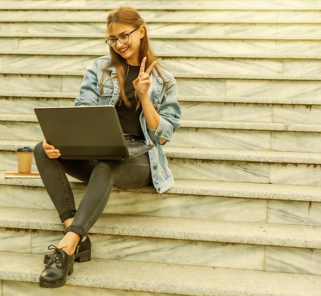 Llamada online. Mujer joven inconformista en chaqueta de mezclilla se comunica con amigos en una conferencia en línea con una computadora portátil mientras está sentado en las escaleras de la ciudad