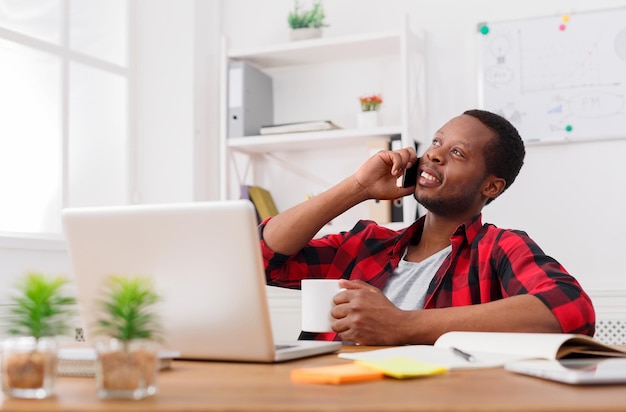 Llamada satisfactoria. Joven empresario negro tiene conversación por teléfono móvil mientras trabaja con una computadora portátil en el interior de la oficina blanca moderna, espacio de copia